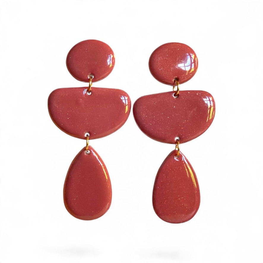 Coloris bois de rose des boucles d’oreilles LOUMA, création artisanale en pâte polymère aux trois formes géométriques suspendues et aux contours doux.